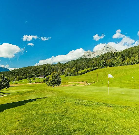 A golf course with a flag on the green, surrounded by trees and mountains under a blue sky with clouds, creating a peaceful and vibrant landscape.