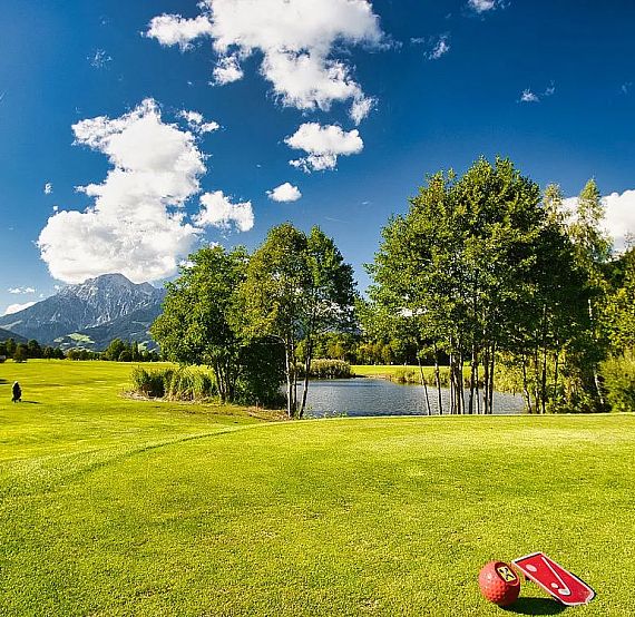 A golf course with lush green grass, a pond, and trees, set against distant mountains under a clear blue sky. Golf equipment lies in the foreground.