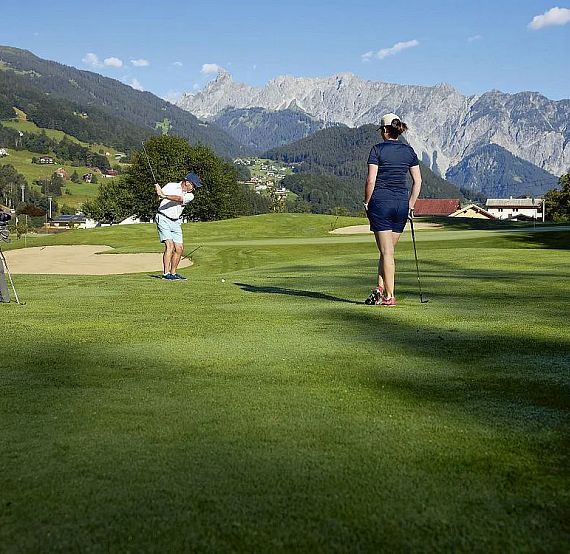 Two golfers on a lush green golf course surrounded by mountains under a clear blue sky. One golfer is about to swing, while the other stands nearby.