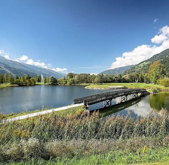 A tranquil lake with a wooden bridge, surrounded by green hills and trees under a blue sky with clouds, creating a peaceful natural scene.